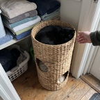 Black cat curled in the top level of the woven rattan cat tower in a utility corner - lifestyle v2