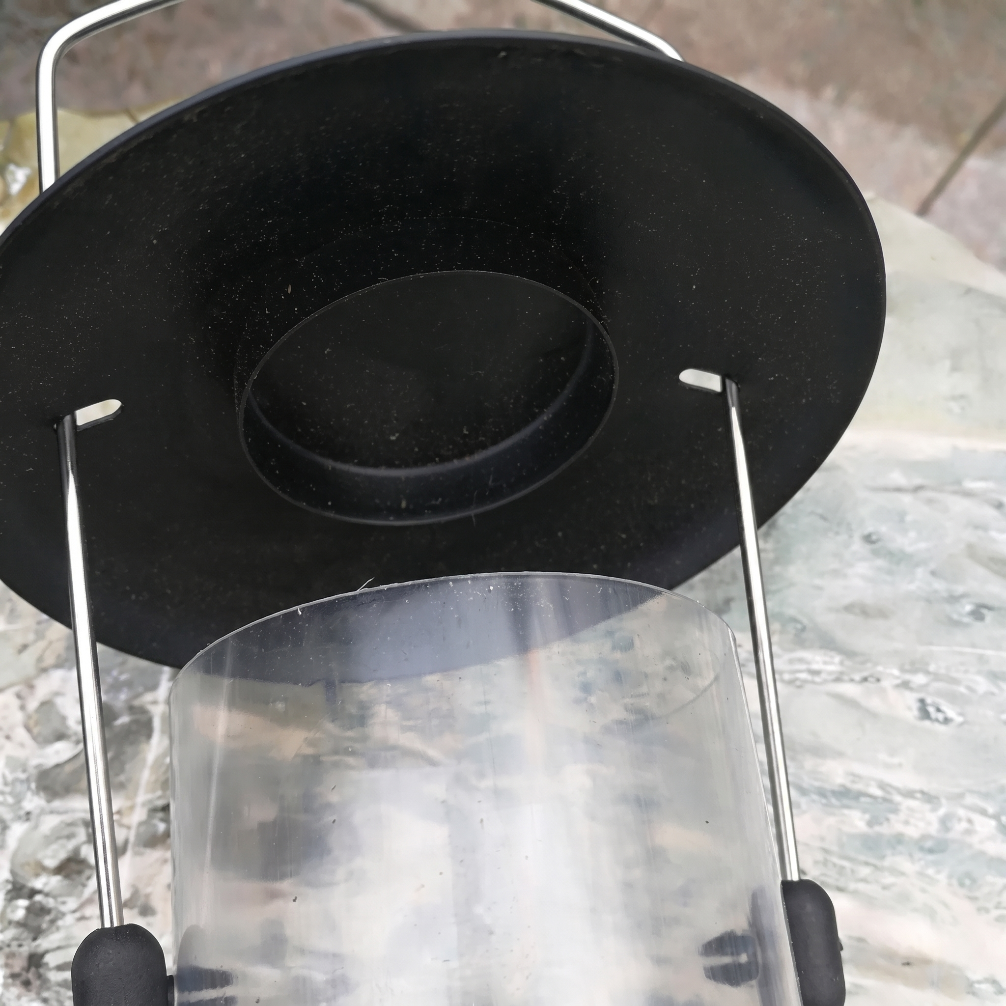 A close-up of a black metal lantern or light fixture with a clear cylindrical glass shade and thin silver metal support arms, photographed outdoors on a stone surface.