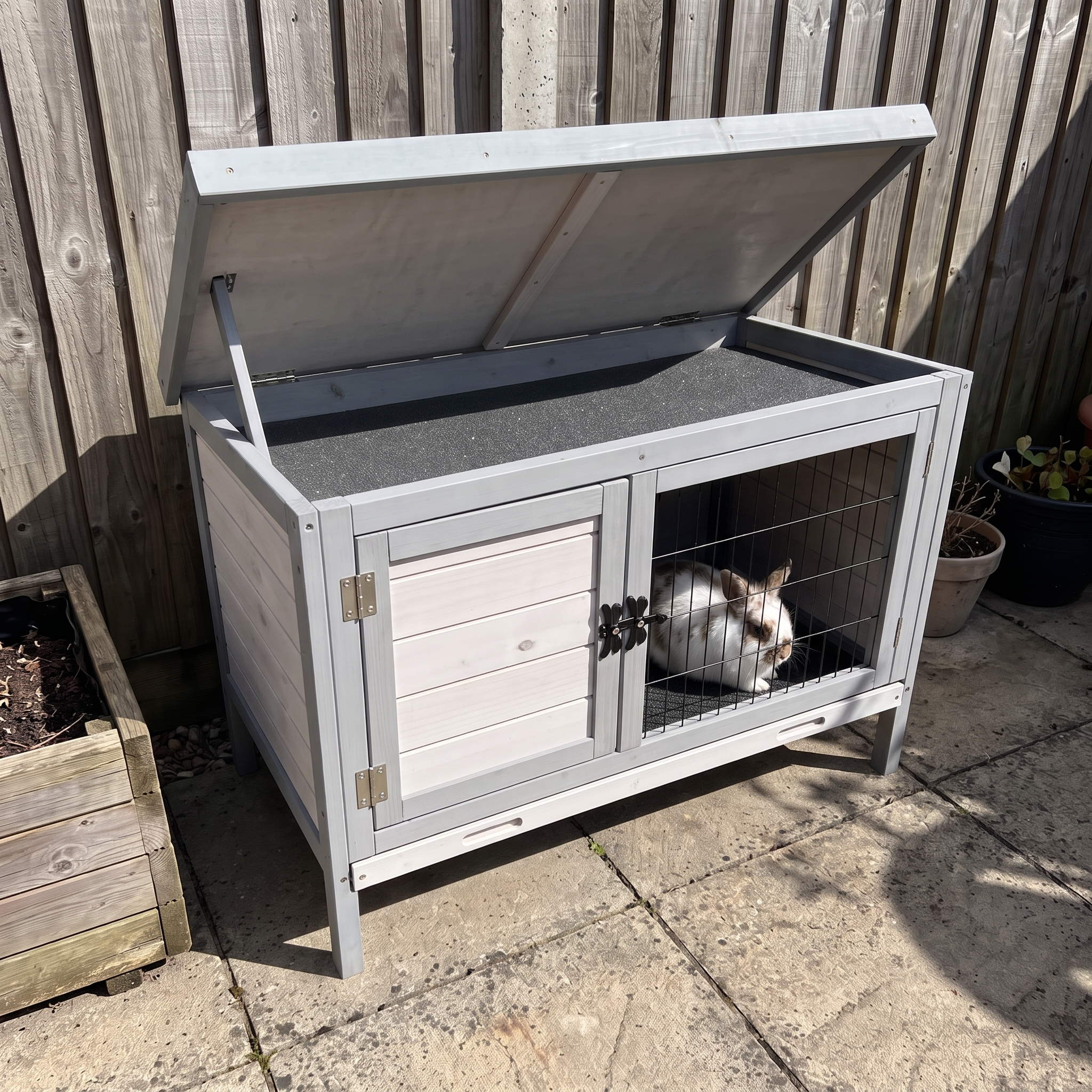 White rabbit sits inside the raised wooden hutch on a patio, lid propped open.
