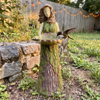 Fairy statue holds a leaf shaped feeder bowl with a bird at its rim in a garden.