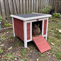 A small rabbit hutch with a red and white wooden exterior, a dark gray roof, and a red ramp, shown outdoors in a garden with a rabbit sitting inside.