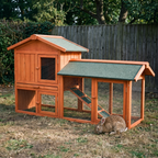 Wooden two-tier rabbit hutch with upper house, mesh run and ramp on a backyard lawn, a brown rabbit grazing in front of the run