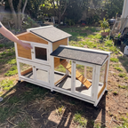 A wooden and white pet playhouse with a small house, wire mesh windows, ramp, and enclosed outdoor area, photographed in a backyard with a person's hand pointing at it.